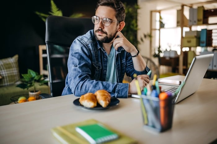 Man with glasses using laptop looks away appearing to be in thought.