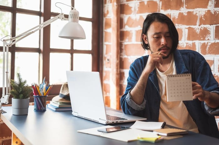 Young man pausing from using his laptop to look at a calendar.