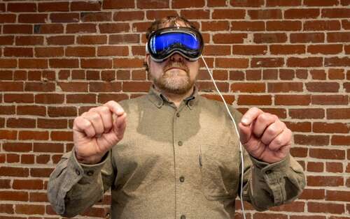 A man wearing an Apple Vision Pro headset holds his hands in front of him, with index fingers pressed against thumbs to control the software