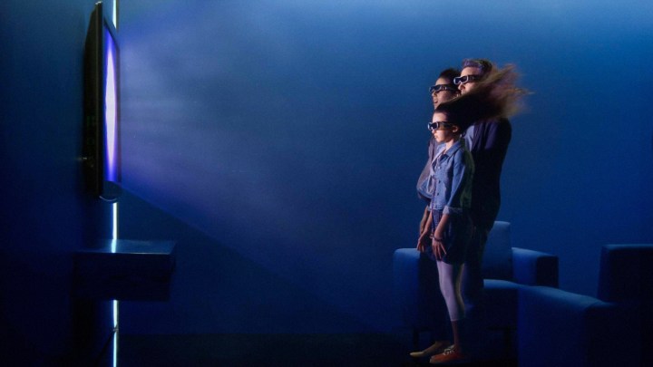 A promotional image showing a child and two adults having their hair blown pack by the power of a 3D TV image. 