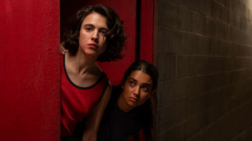 Margaret Qualley and Geraldine Viswanathan peer around a corner in a still from Drive Away Dolls