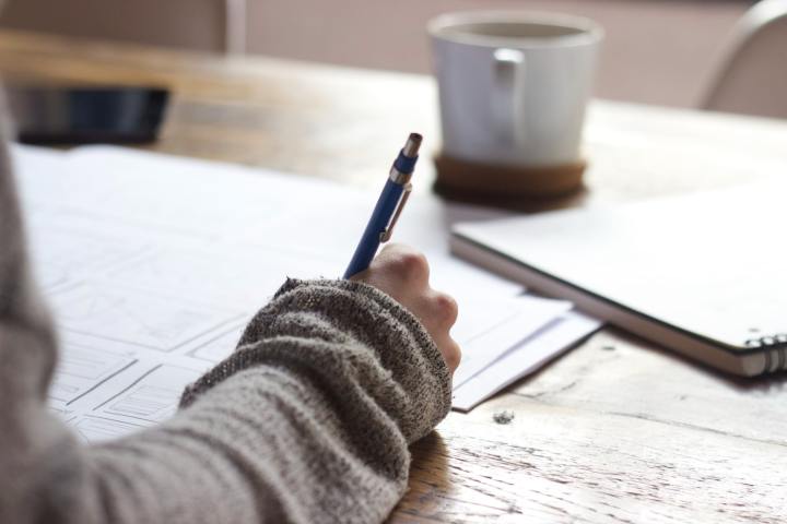 A person studying and writing notes onto a piece of paper. There is a coffee cup and binder in the distance.