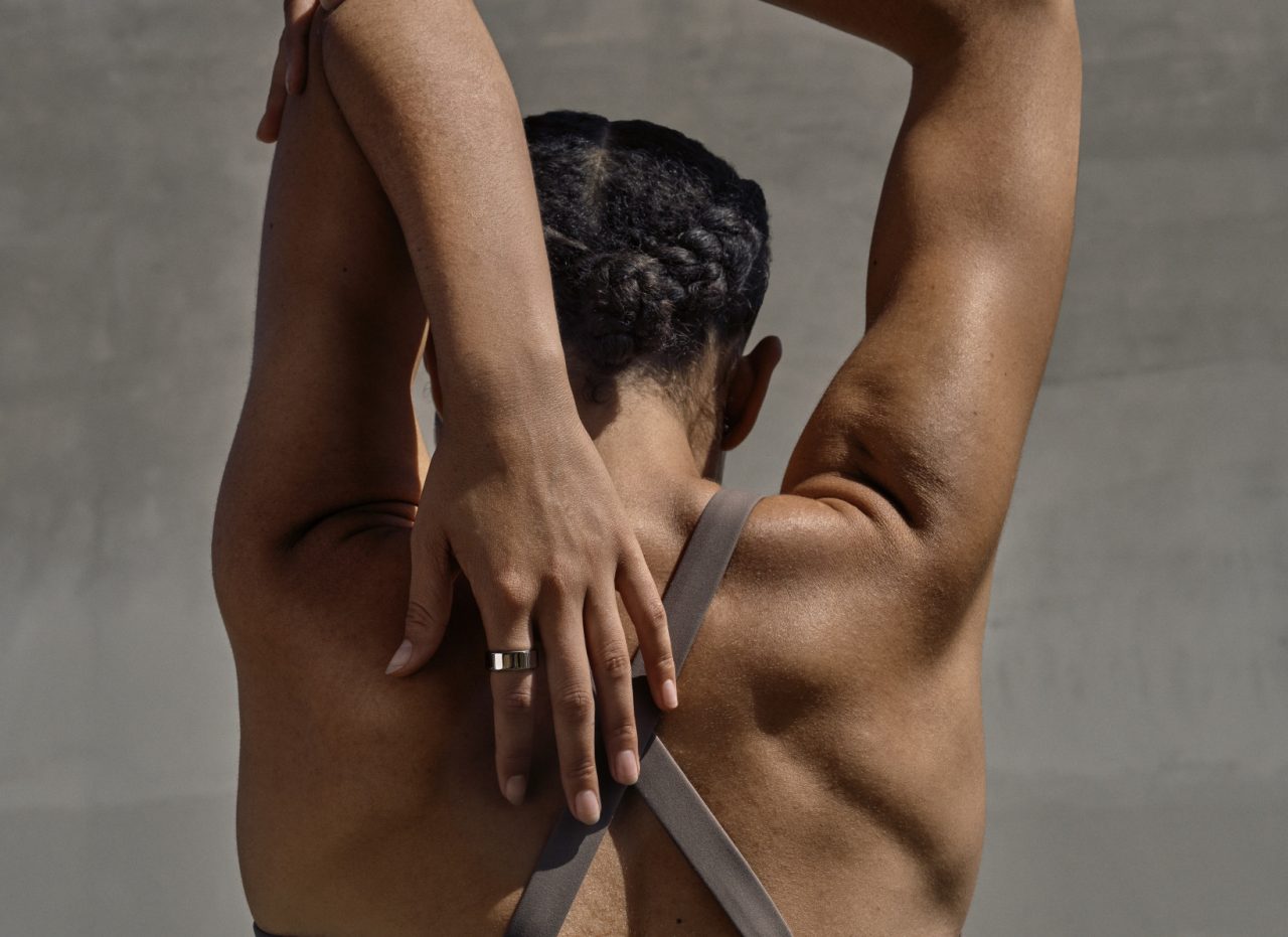 A woman wearing a sports bra is seen from behind stretching her tricep and wearing an Oura smart ring health tracking wearable on her finger