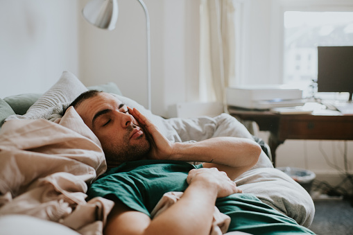 A man wakes up on a sofa, surrounded by sheets and duvets and pillows. He rubs his eyes and face as he tries to rouse himself.