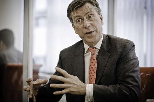 A man in a grey suit, striped shirt, and pink tie gestures with his hands while sitting in a corporate room.