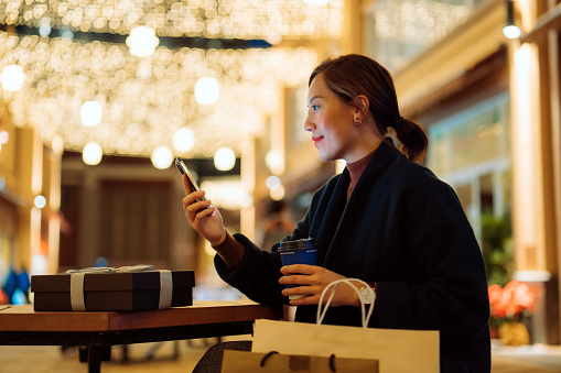 Beautiful young Asian woman relaxing on a bench and checking smartphone after shopping in the city, with decorated high street and illuminated Christmas lights in background. Enjoying Christmas shopping. Festive vibes. Christmas is almost here