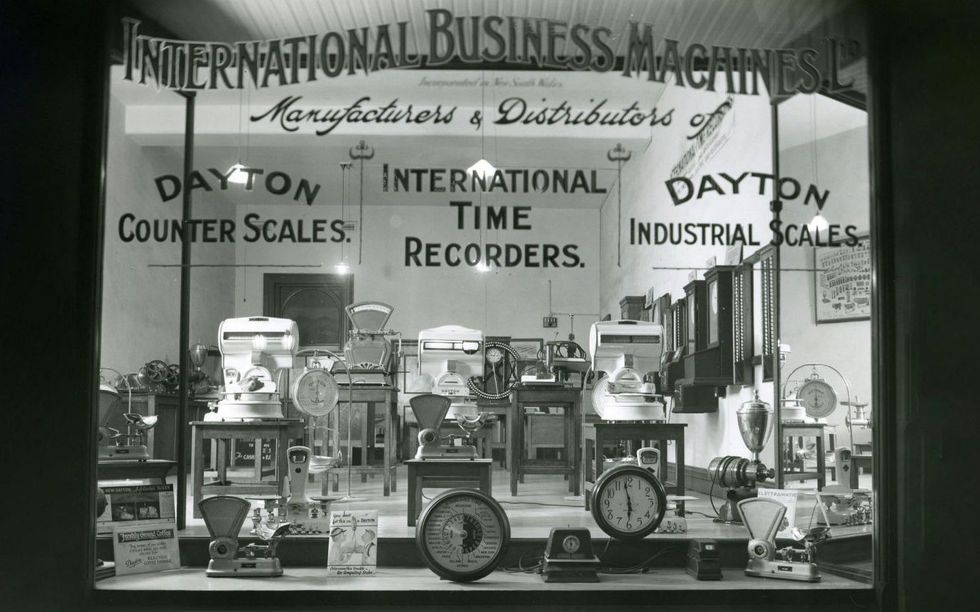 Black and white photo of a shop window displaying various scales and clocks. Across the top, it reads International Business Machines. 