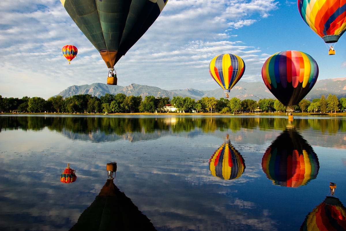 Colorful hot air balloons glide over Prospect Lake in Colorado Springs, CO, at the Colorado Balloon Classic; Cheyenne Mountain is in the background.