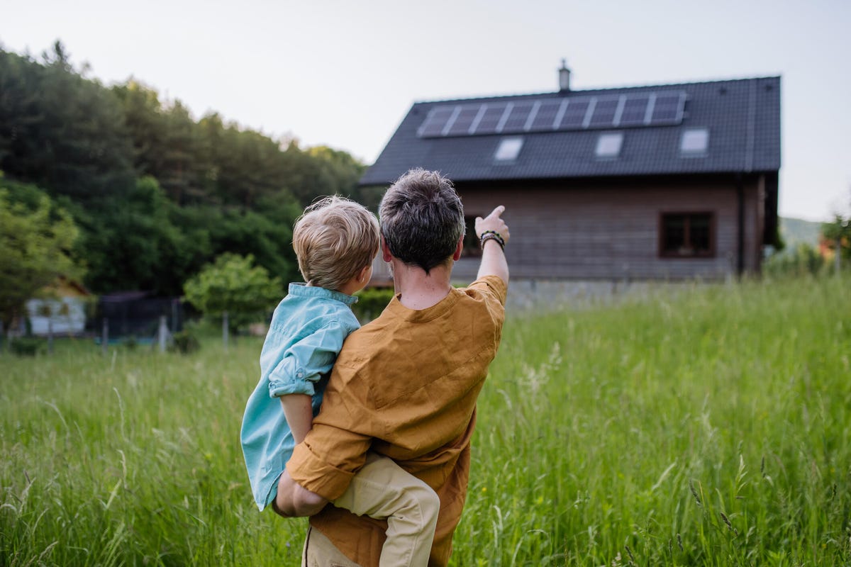 A father holds a young child while pointing at a house with solar panels on it.