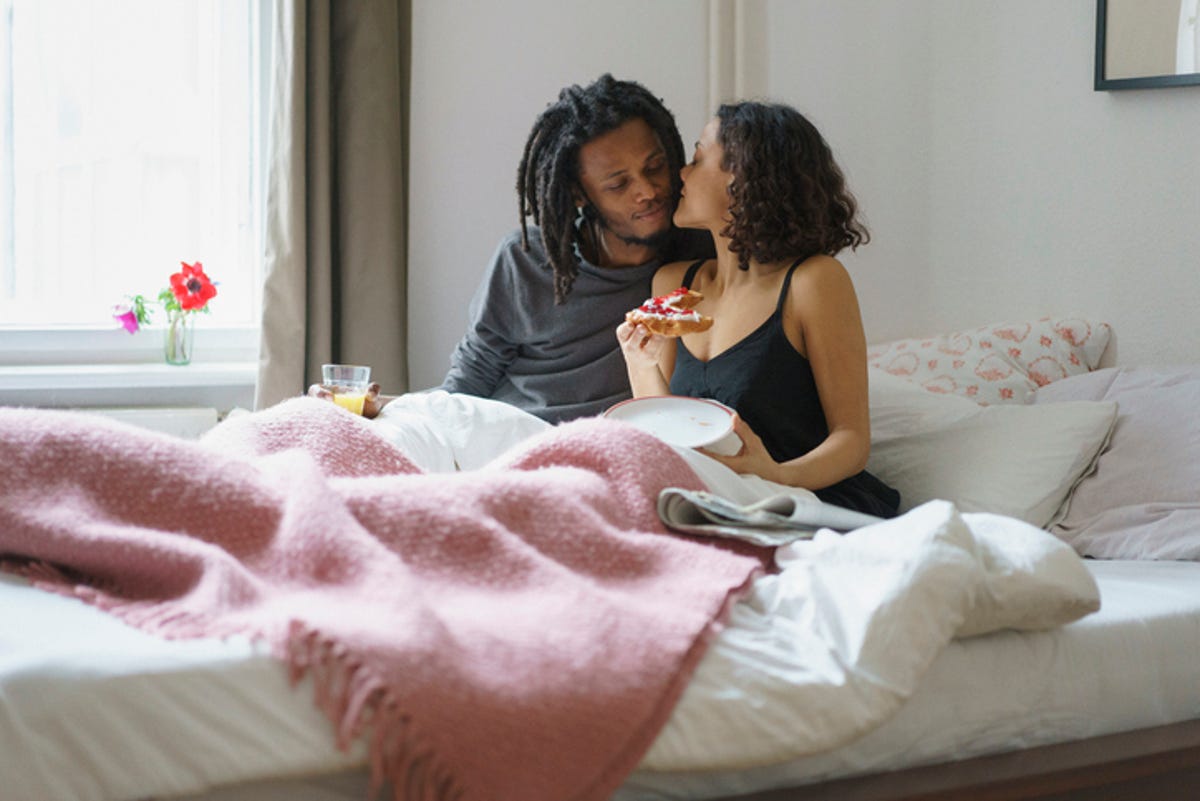 Young woman whispering to a man while holding food in bed