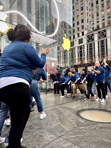 Apple employees in dark blue shirts in front of the Apple store stretch their hands out in anticipation.