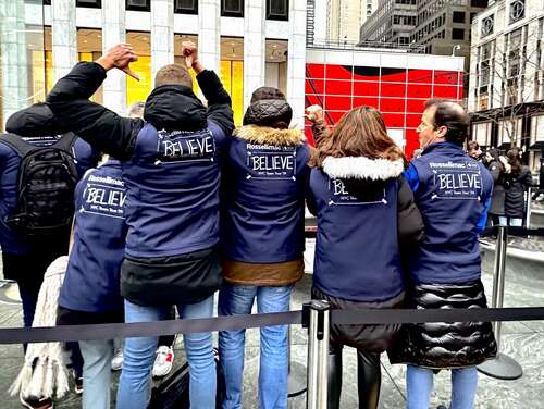 Five people stand in a line with their backs to the camera. They are all wearing the same vests.