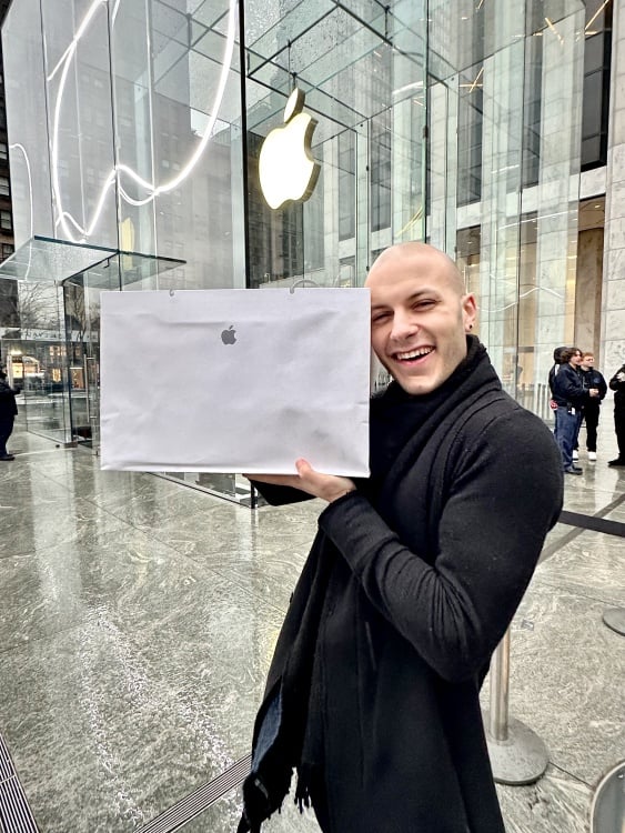 A white man in a black ensemble olds an Apple bag up to his head in front the the 5th avenue apple store.