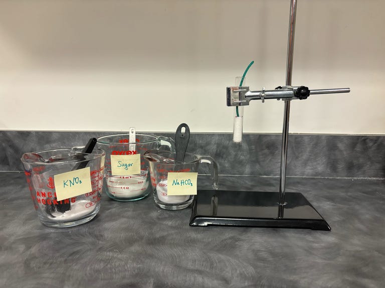 Glass measuring cups containing potassium nitrate, sugar, and baking soda sit beside a test tube containing a mixture of the three, plus a fuse.