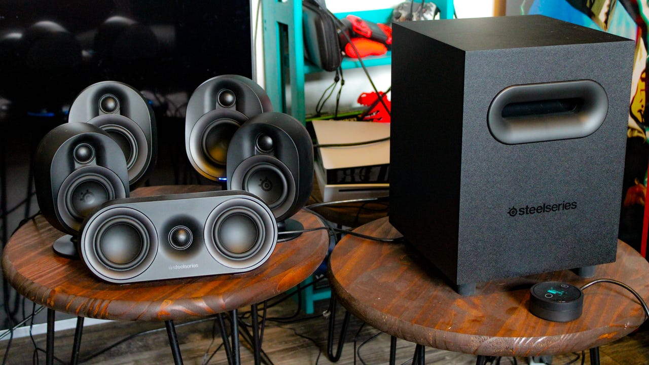 A pair of Arena 9 speakers on a table that is between two chairs. An orange dinosaur decor piece is between the speakers, and a cat sits above them in a window seat, looking at the camera.