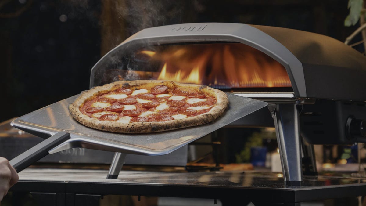 Someone uses an Ooni pizza peel to slide a pepperoni pizza out of an Ooni pizza oven set up on a metal tabletop.