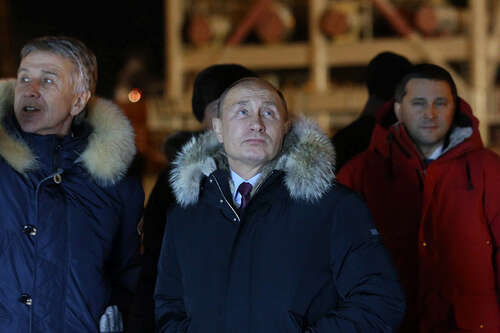 Three men stand in front of a large steel tower, at night, wearing heavy parkas. A man on the left gestures at something behind the camera, while Vladimir Putin stands in the middle, looking on with an expression of interest.