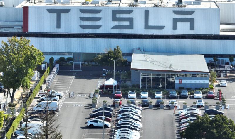 Aerial view of a Tesla factory shows a giant Tesla logo on the side of the building, and a parking lot filled with cars.