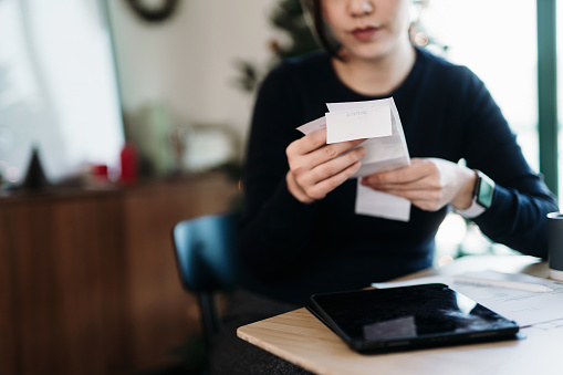 Young Asian woman managing personal banking and finance. Planning budget and calculating expenses with digital tablet while going through various expense receipts at home after shopping. Home finances. Home budgeting. Banking and finance concept