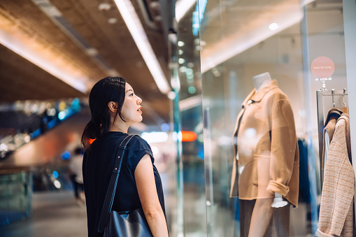 Young Asian woman standing outside a boutique looking at window display while shopping in shopping mall. Window shopping. Sale season