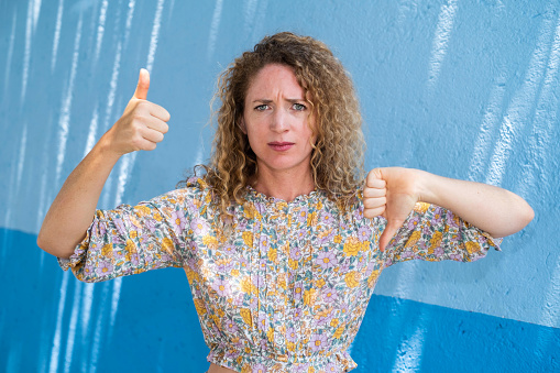 Young woman with curly blonde hair looking at the camera confused and giving a thumbs up and thumbs down