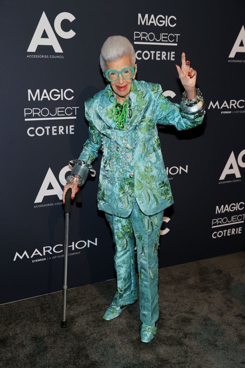 A person waves while wearing a flamboyant turquoise and holding a cane.