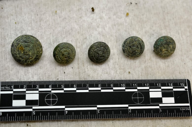 Five weathered buttons are shown against a ruler, indicating they are only a few inches wide. Some clearly show "16th Battalion Canadian Scottish Regiment."