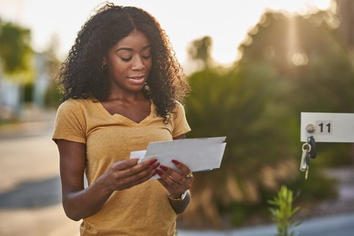 A woman stands in front of a multi-unit mailbox that she opened with a key and looks through the mail she received.