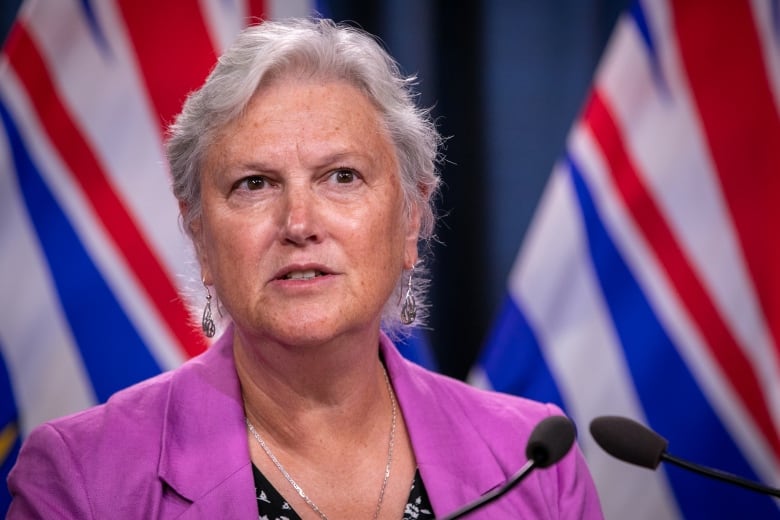 A white woman with white hair and wearing a pink blazer stands in front of two B.C. flags at a podium.