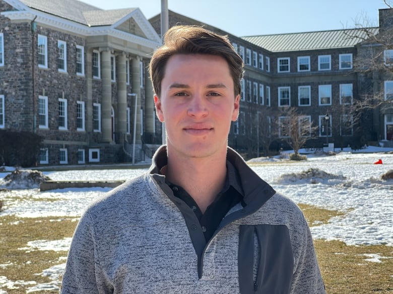 A clean shaven young man with brown hair looks at the camera. Behind him are brick buildings on a university campus.