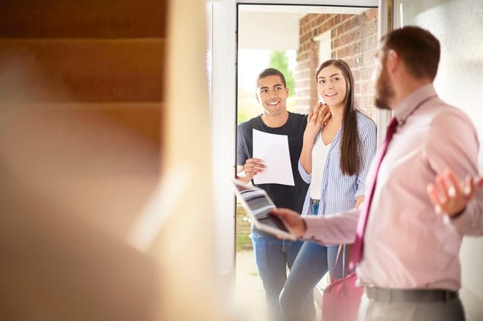 A man and woman looking through the door of a house held open by a realtor.