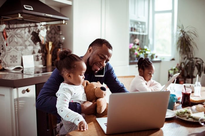 A dad making a phone call and looking at his laptop while sitting at the kitchen table having breakfast with his two young daughters.