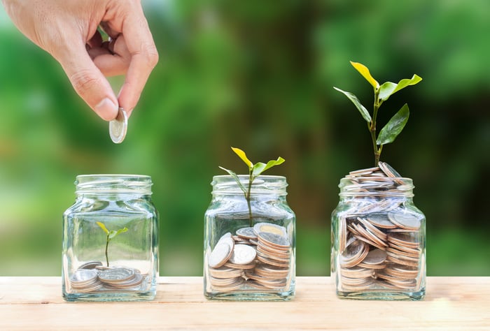 A person stacking coins into glass jars that sprout larger plants the more coins fill the jar illustrates the concept of compounding wealth over time. 