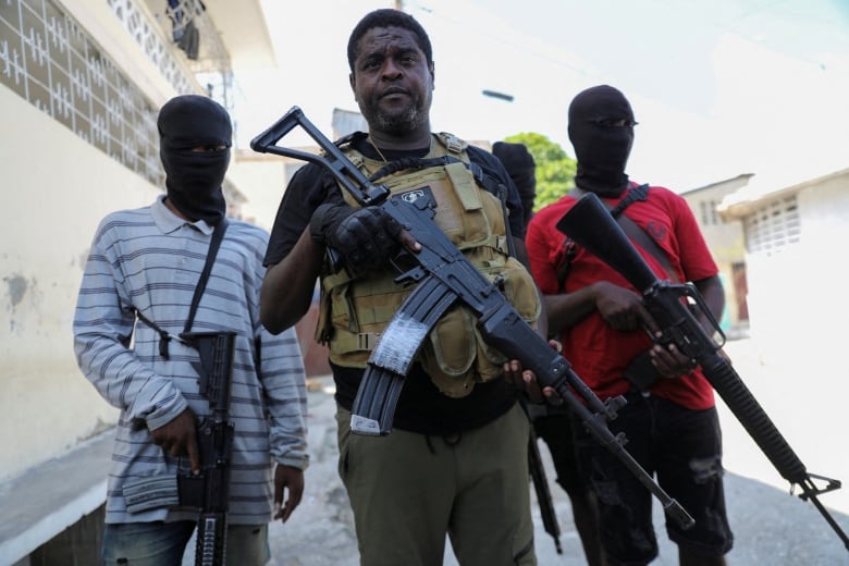 Three men with large guns are seen standing in an outdoor setting. The man in the middle is clean shaven, dark complected. The other two men wear masks concealing their identity.