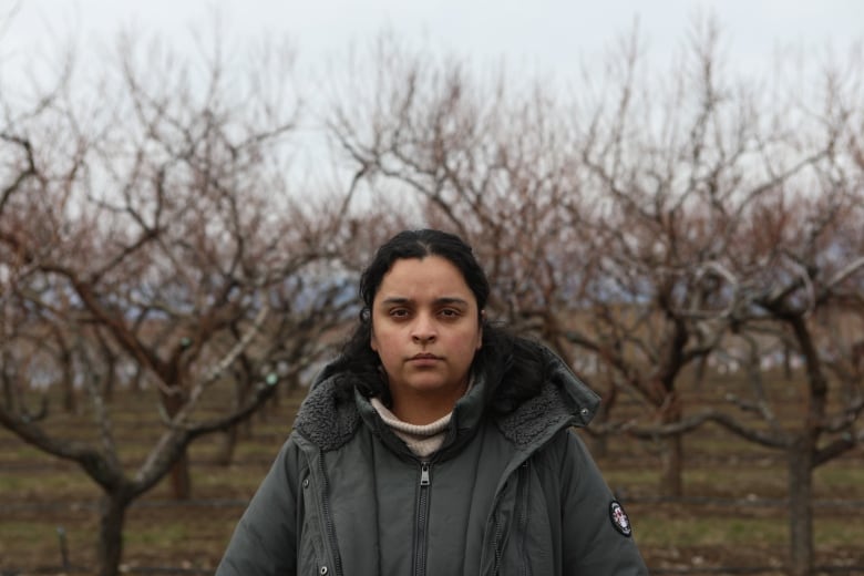 A woman stands in a barren orchard with a serious look on her face.