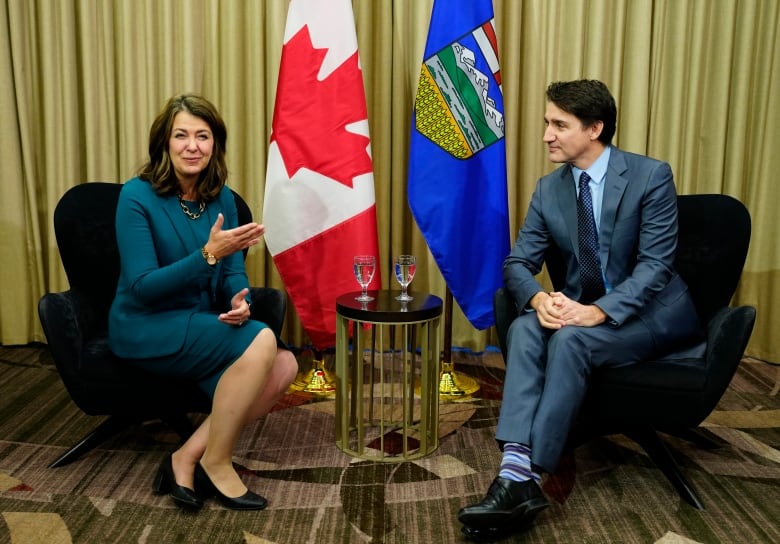 Prime Minister Justin Trudeau seated opposite Alberta Premier Danielle Smith. A small side table with water glasses sits between them with flags of Alberta and Canada on poles in the background.