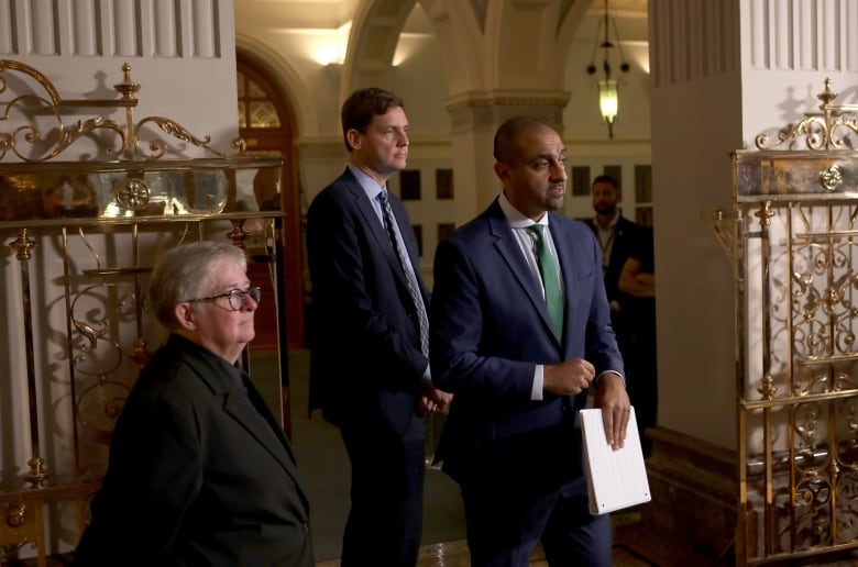 A white middle-aged woman stands with hands held behind her back. She wears a black suit and black-rimmed glasses. On her left, stands a tall white middle-aged man wearing a blue suit and to his left stands a South Asian man, also tall, wearing a navy blue suit and a green tie.