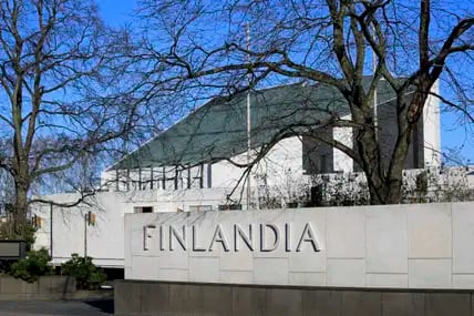 Blick auf die weiße Finlandia Hall mit grünem Dach in Helsinki, deren Name in Großbuchstaben an einer Mauer steht, davor Bäume