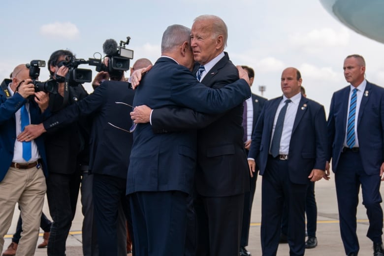 Two older men in suits embrace on an airport tarmac as others look on.