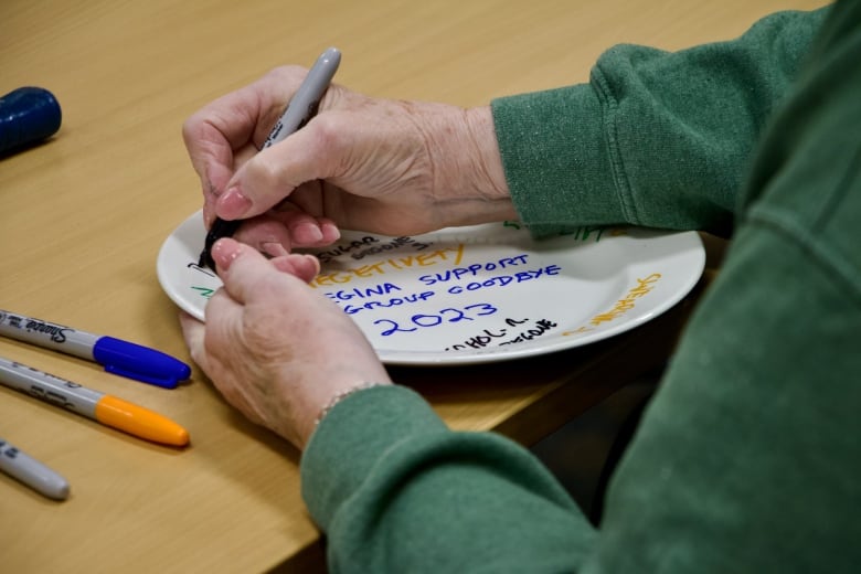 A woman's hands writing on a white plate using a permanent marker. 'Regina support group goodbye 2023' can be seen on the plate.