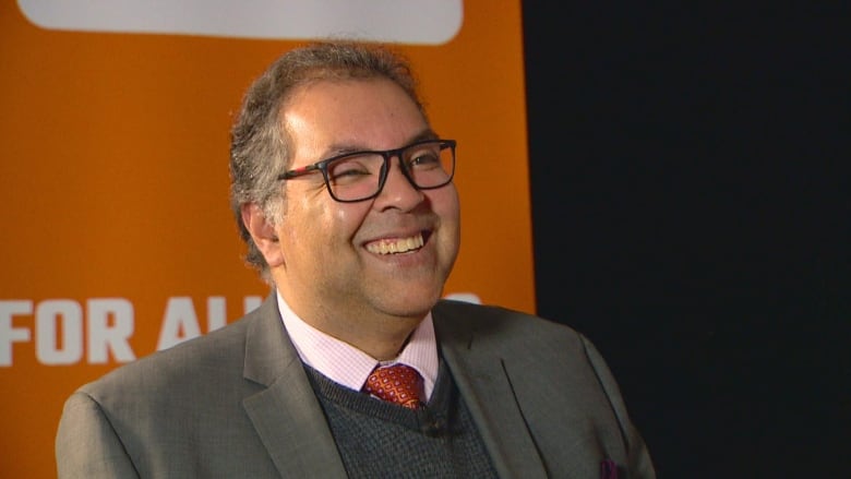 A man wearing a suit smiles in front of a backdrop.