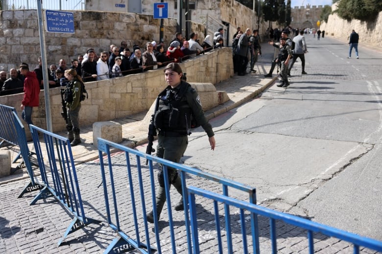 A woman in a police-type uniform is shown near a barricade as behind her a lineup of people is shown.