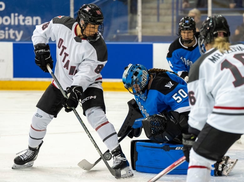 A hockey player looks at the net and opposing goaltender behind her. She doesn't see the puck between her skates.