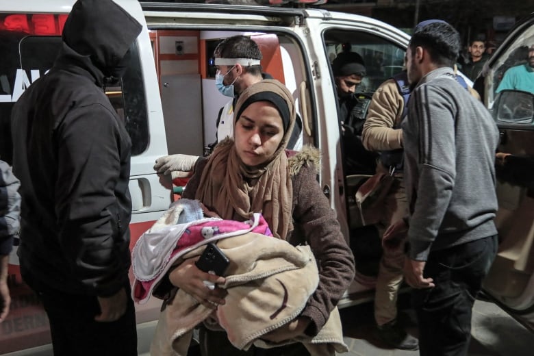 A woman in a headscarf is shown in the foreground carrying a covered baby in her arms. An ambulance and several others are shown in the background.