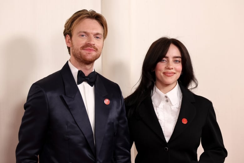 A man and a woman wearing suits smile. Both are wearing red pins on their lapels.