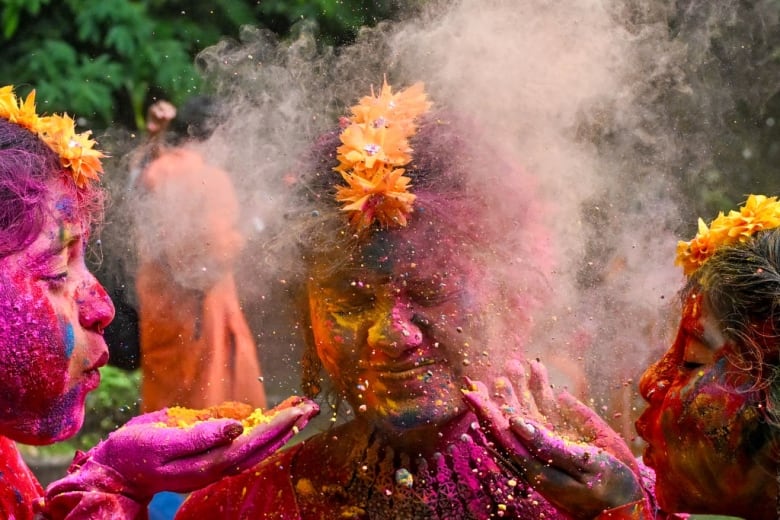 Two women blow coloured powder on the face of a third.