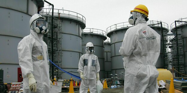 Drei Menschen in Schutzanzügen vor H4-Wassertanks mit radioaktiv belastetem Wasser