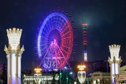 Blick bei Nacht auf das blau-rosa erleuchtete Riesenrad Sun Ferris Wheel in Moskau, zu beiden Seiten erleuchtete Türme