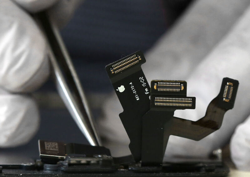 Cables emanating from an iPhone under repair, with gloves hands holding a tweezer over the phone