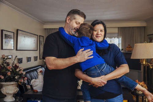 A photo of a couple, wearing black shirts, and their child, with arms slung over both parents' shoulders